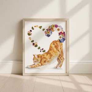 May include: A framed art print featuring an orange tabby cat stretching, with a heart-shaped floral tail. The heart is composed of various dried flowers and greenery. The artwork is set against a white background, framed in light wood.
