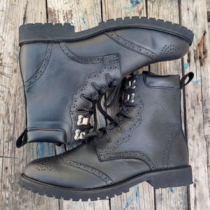 May include: Black leather boots with decorative perforations and scalloped edges. The boots have a lace-up design with metal eyelets and a sturdy, lug sole. The boots are shown on a wooden surface.