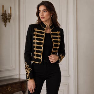 Pode incluir: Uma jaqueta preta de estilo militar com gola alta, mangas compridas e detalhes de cordão trançado em tom dourado na frente. A jaqueta inclui botões dourados e padrões de bordado dourado intrincados nos punhos e nos painéis frontais.