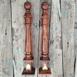 May include: A pair of wooden balusters, stained a deep reddish-brown. Each has a spherical top, a turned central section, and a square base with a gold-coloured inlay. The wood grain is visible, enhancing the natural appearance. The balusters are likely for a staircase.