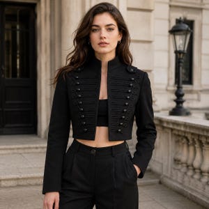 May include: A black cropped military-style jacket featuring a high collar, long sleeves, and a front closure adorned with horizontal braided cord trim and matching black buttons. The jacket is worn over a black crop top and high-waisted black trousers.