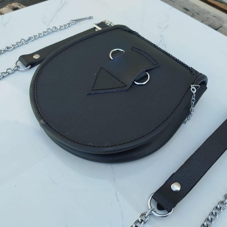 Pu&ograve; includere: Una borsa in pelle nera con una tracolla a catena argentata e una chiusura a lembo. La borsa ha una forma triangolare sul davanti e due anelli argentati.