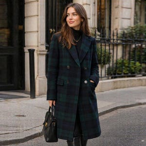 May include: A double-breasted, long coat in a green and navy plaid pattern. The coat has a classic lapel, two rows of buttons, and pockets. The person is also wearing a black turtleneck and carrying a black handbag.