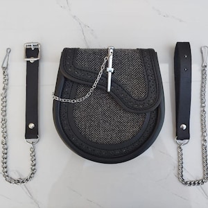 May include: A black leather sporran with a silver chain and a silver kilt pin. The sporran has a herringbone pattern and a black leather strap.