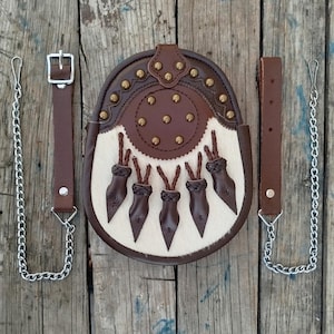 Op de afbeelding: Een bruine leren sporran met een witte bontklep, versierd met messing studs en kwastjes. Het wordt geleverd met een bruine leren riem en een zilveren ketting.