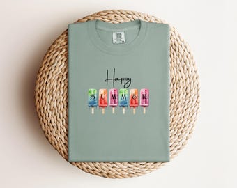 May include: A sage green t-shirt featuring the text "Happy" in black script above six colorful watercolor-style popsicle illustrations. Each popsicle contains a single black letter spelling "SUMMER" and shows a melting effect at the bottom.