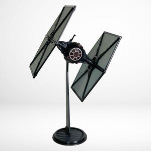May include: A black TIE fighter model with a stand. The TIE fighter has two wings that are angled upwards. The stand is black and has a round base.