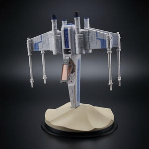 May include: A white and blue starfighter model with four extended wings, laser cannons, and a cockpit with a tinted orange canopy. The spacecraft is mounted on a tan, textured base that resembles a sand dune, set against a dark background.