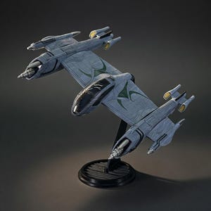 May include: A detailed model of a futuristic spacecraft, primarily gray with green and yellow accents. The model is mounted on a black stand, showcasing its intricate design and aerodynamic shape. The spacecraft has multiple wings and engine components.