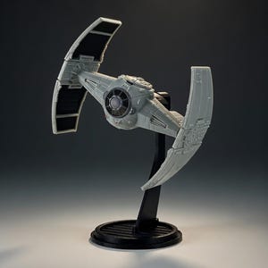 May include: A gray model of a TIE fighter spacecraft, a fictional starship from a science fiction franchise. The model is mounted on a black stand with a circular base. The spacecraft has two large solar panels and a central cockpit.