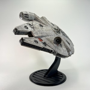 May include: A detailed model of the Millennium Falcon starship, in shades of gray with red accents, mounted on a black stand. The model features intricate paneling and a circular radar dish. The stand has a round base.