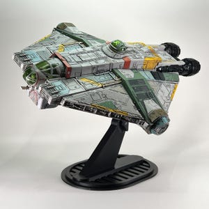 May include: A detailed model of a spacecraft, featuring a color scheme of gray, green, yellow, and red. The model is mounted on a black stand with an oval base. The spacecraft has intricate paneling and design elements, with visible engines and a cockpit.