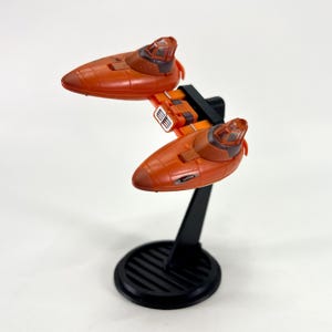 May include: A miniature orange spacecraft model with two separate cockpit sections. The model is mounted on a black stand with a circular base. The spacecraft has a sleek, aerodynamic design, with detailed cockpit windows and engine components.