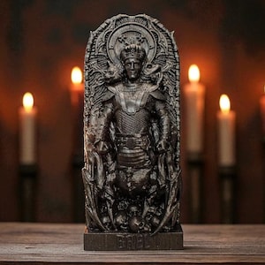 May include: A dark gray statue of a crowned figure in armor, set against a blurred background of lit candles. The statue is intricately detailed, with a base that reads "GRIEL". The scene is set on a wooden surface.