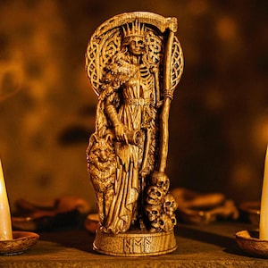 Puede incluir: Una detallada estatua dorada de una figura vestida con una túnica que sostiene una guadaña, con un lobo y calaveras en la base. Dos velas encendidas flanquean la estatua, creando un ambiente cálido y místico. La estatua tiene intrincadas tallas.