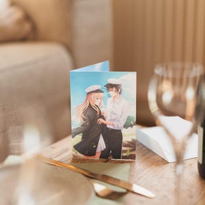 May include: A greeting card featuring a cartoon illustration of a couple in sailor outfits. The woman has blonde hair and is wearing a black coat and a white hat. The man has black hair and is wearing a white shirt and a black hat. The background is a blue sky with white clouds.