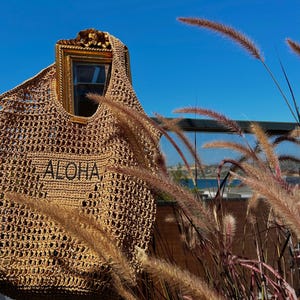 Op de afbeelding: Een bruine gehaakte draagtas met een gouden frame en het woord "ALOHA" geborduurd op de voorkant. De tas hangt aan een hek met hoog gras op de achtergrond.