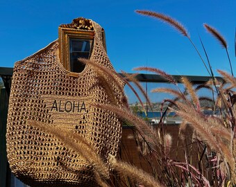 May include: A brown crocheted tote bag with a gold frame and the word "ALOHA" embroidered on the front. The bag is hanging on a fence with tall grass in the background.