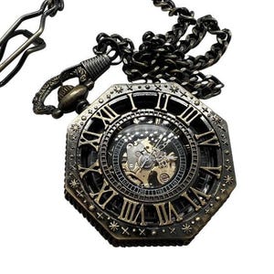 Puede incluir: Un reloj de bolsillo de estilo vintage con una cara octogonal y marcas de números romanos. El reloj tiene un acabado dorado y una cadena negra.