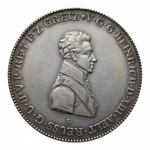 May include: A silver coin featuring a profile portrait of a man in a military-style jacket. The coin has a detailed edge and is inscribed with text around the perimeter. The coin's design is centered on a white background.