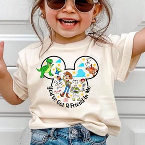 May include: A cream-colored t-shirt featuring a graphic of Disney's Toy Story characters inside a Mickey Mouse head shape. The text "You've Got A Friend In Me" is printed around the design. The shirt is worn by a child.