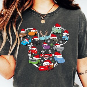 May include: Dark gray t-shirt featuring a design of cartoon cars wearing Santa hats, arranged in a Mickey Mouse head shape. The word "Cars" is printed in white. The shirt is worn with blue jeans and white sneakers.