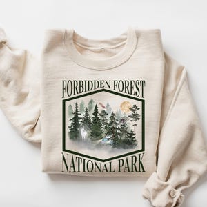 May include: A cream-colored sweatshirt with the words "FORBIDDEN FOREST NATIONAL PARK" in a black, stylized font. The graphic features a forest scene with trees, mountains, and a full moon. The sweatshirt is folded with the sleeves rolled up.