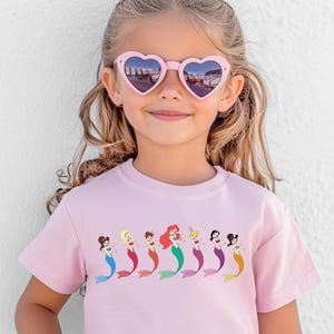 May include: A light pink t-shirt featuring a row of cartoon mermaids with colorful tails. The mermaids have different hair colors and styles. The girl in the image is wearing pink heart-shaped sunglasses.