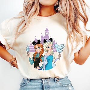 May include: Cream-colored t-shirt featuring a cartoon illustration of two princesses with mouse ears in front of a purple castle. One princess holds ice cream, the other cotton candy. The princesses wear blue and black outfits.