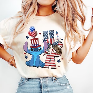 K&ouml;nnte beinhalten: Cremefarbenes T-Shirt mit einer blauen Cartoonfigur, die einen patriotischen Hut tr&auml;gt, und dem Text "USA" in Rot, Wei&szlig; und Blau. Das Design umfasst Sterne, Feuerwerk und ein Leopardenmuster. Das Shirt wird mit hellblauen Jeans getragen.