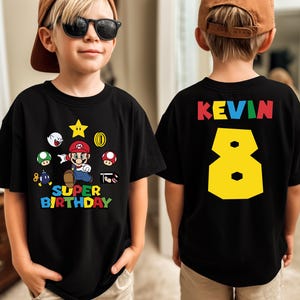 K&ouml;nnte beinhalten: Schwarzes T-Shirt mit einem Super Mario-Motiv und dem Text "SUPER BIRTHDAY". Auf der R&uuml;ckseite stehen der Name "KEVIN" und die gelbe Zahl "8". Das Kind tr&auml;gt eine braune Baseballkappe und eine Sonnenbrille. F&uuml;r den 8. Geburtstag.