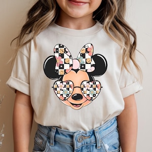Puede incluir: Una camiseta de manga corta color crema con un gr&aacute;fico de una cabeza de rat&oacute;n de dibujos animados que lleva gafas en forma de coraz&oacute;n. El lazo y los cristales de las gafas tienen un patr&oacute;n de tablero de ajedrez blanco y negro, flores rosas y caras sonrientes amarillas.