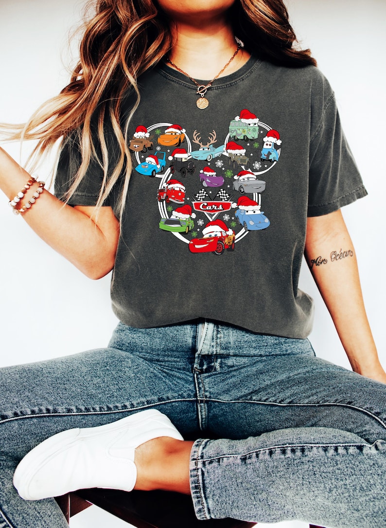 May include: Dark gray t-shirt featuring a design of cartoon cars wearing Santa hats, arranged in a Mickey Mouse head shape. The word "Cars" is printed in white. The shirt is worn with blue jeans and white sneakers.