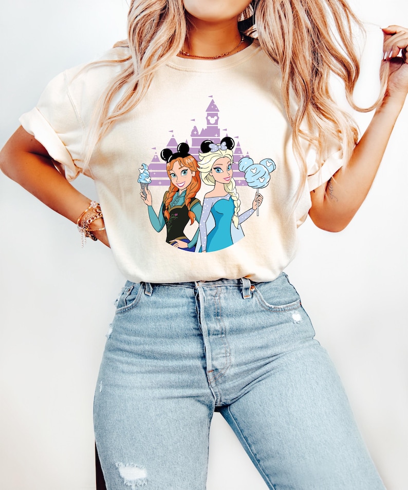 May include: Cream-colored t-shirt featuring a cartoon illustration of two princesses with mouse ears in front of a purple castle. One princess holds ice cream, the other cotton candy. The princesses wear blue and black outfits.