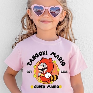 May include: A light pink t-shirt featuring a cartoon image of Tanooki Mario, with the text "Tanooki Mario" and "EST 1985". The shirt is worn by a young person wearing pink heart-shaped sunglasses.