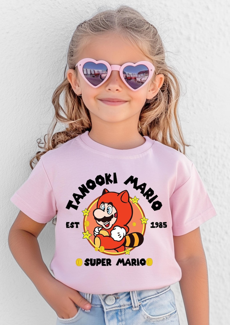 May include: A light pink t-shirt featuring a cartoon image of Tanooki Mario, with the text "Tanooki Mario" and "EST 1985". The shirt is worn by a young person wearing pink heart-shaped sunglasses.