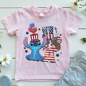 K&ouml;nnte beinhalten: Ein hellrosa T-Shirt mit einer blauen Cartoon-Figur, die einen patriotischen Hut tr&auml;gt und Luftballons h&auml;lt. Das Shirt hat den Text "USA" in einem Design mit der amerikanischen Flagge und einem Leopardenmuster "S". Rote und blaue Sterne und ein Feuerwerk vervollst&auml;ndigen das Design.
