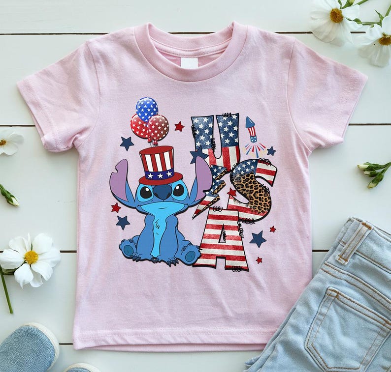 K&ouml;nnte beinhalten: Ein hellrosa T-Shirt mit einer blauen Cartoon-Figur, die einen patriotischen Hut tr&auml;gt und Luftballons h&auml;lt. Das Shirt hat den Text "USA" in einem Design mit der amerikanischen Flagge und einem Leopardenmuster "S". Rote und blaue Sterne und ein Feuerwerk vervollst&auml;ndigen das Design.