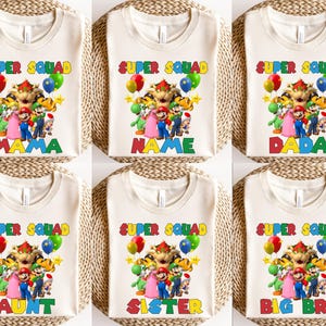 K&ouml;nnte beinhalten: Cremefarbene T-Shirts mit einer farbenfrohen Grafik mit Charakteren aus der Super Mario Bros. Franchise. Jedes Shirt hat die Worte "SUPER SQUAD" &uuml;ber der Grafik und eine andere Familienrolle darunter, wie "MAMA" oder "SISTER".