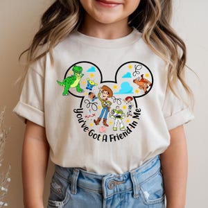 May include: A beige t-shirt featuring a graphic of characters from the animated film Toy Story. The design includes Woody, Buzz Lightyear, Rex, and other characters within a Mickey Mouse-shaped outline, with the text "You've Got A Friend In Me."