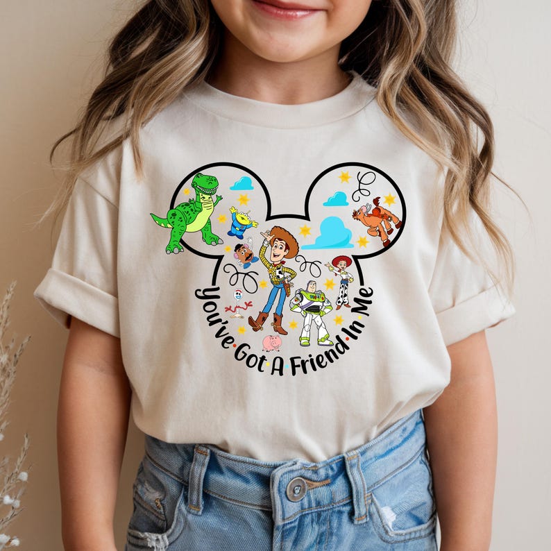 May include: A beige t-shirt featuring a graphic of characters from the animated film Toy Story. The design includes Woody, Buzz Lightyear, Rex, and other characters within a Mickey Mouse-shaped outline, with the text "You've Got A Friend In Me."