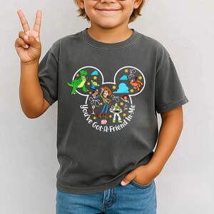 May include: Dark gray t-shirt featuring a colorful graphic of Toy Story characters inside a Mickey Mouse head shape. The text "You've Got A Friend In Me" is printed in a circular pattern around the design. A child is wearing the shirt.