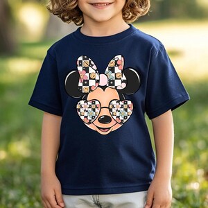 Puede incluir: Una camiseta azul marino de manga corta con un gr&aacute;fico de una cabeza de rat&oacute;n de dibujos animados que lleva gafas de sol en forma de coraz&oacute;n y un lazo grande. El lazo y las gafas tienen un patr&oacute;n de tablero de ajedrez blanco y negro, caras sonrientes y flores.