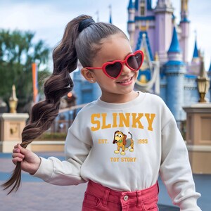 May include: A cream-colored sweatshirt featuring the word "SLINKY" in yellow block letters, with "EST. 1995" and "TOY STORY" below a cartoon image of Slinky Dog. The sweatshirt is worn by a person wearing red heart-shaped sunglasses.