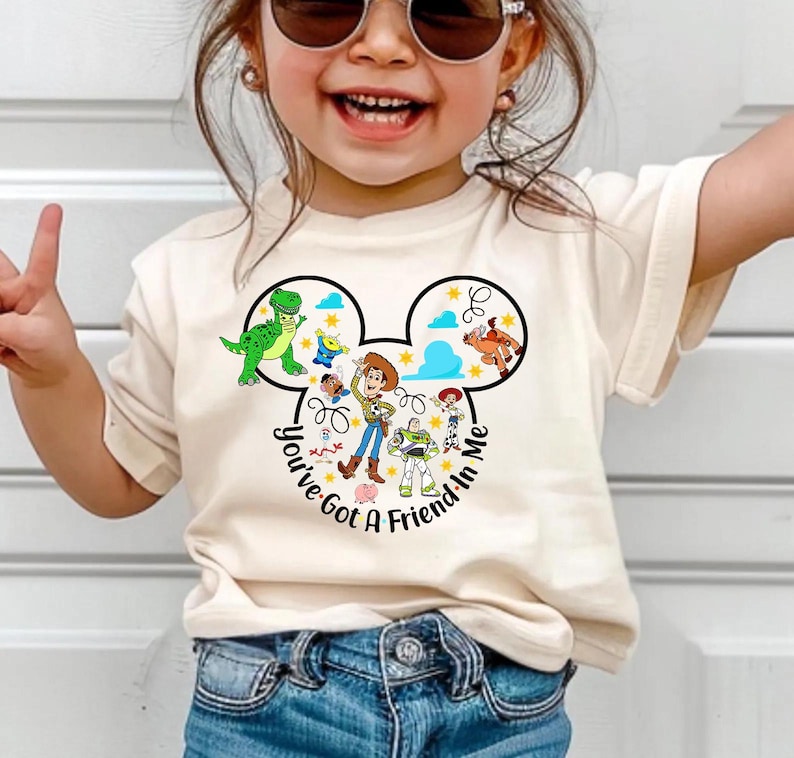 May include: A cream-colored t-shirt featuring a graphic of Disney's Toy Story characters inside a Mickey Mouse head shape. The text "You've Got A Friend In Me" is printed around the design. The shirt is worn by a child.