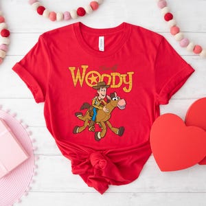 May include: Red t-shirt featuring a cartoon illustration of Woody riding a horse, with the text "Woody" in yellow. The shirt is tied in a knot at the bottom. Decorative hearts and a garland are in the background.