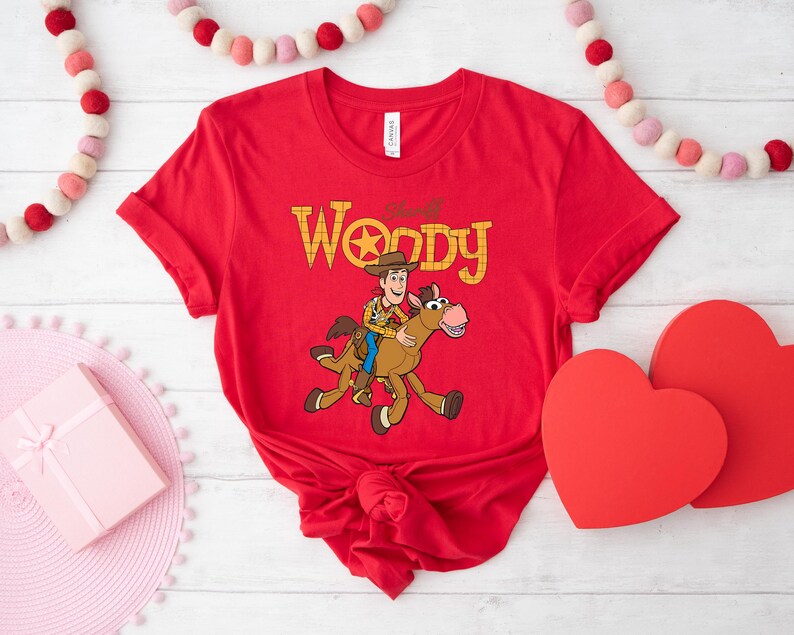 May include: Red t-shirt featuring a cartoon illustration of Woody riding a horse, with the text "Woody" in yellow. The shirt is tied in a knot at the bottom. Decorative hearts and a garland are in the background.