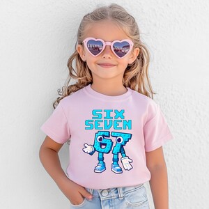May include: A light pink t-shirt with the words "SIX SEVEN" in blue pixelated font. Below the text is a cartoon graphic of the numbers 6 and 7 with arms, legs, and shoes. The child is wearing heart-shaped sunglasses.