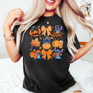 Puede incluir: Camiseta negra con un dise&ntilde;o colorido de Halloween. El dise&ntilde;o incluye calabazas, lazos naranjas, hojas de oto&ntilde;o y un personaje de dibujos animados azul. La camiseta es de estilo casual.
