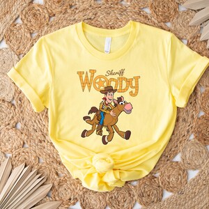 May include: A yellow t-shirt featuring a graphic of Sheriff Woody riding a horse. The text "Sheriff Woody" is above the illustration. The shirt is made of a soft material and has rolled-up sleeves.